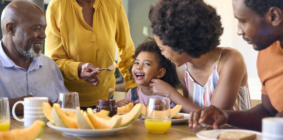 Family Shot With Grandparents Parents And Granddaughter Eating Breakfast Around Table At Home