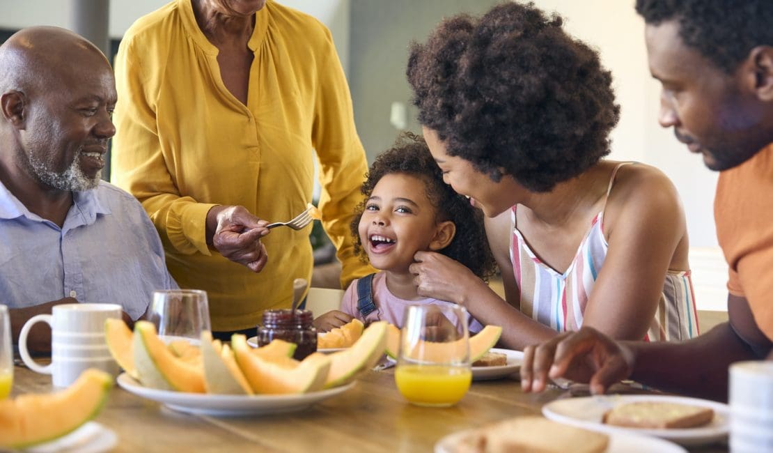 Family Shot With Grandparents Parents And Granddaughter Eating Breakfast Around Table At Home