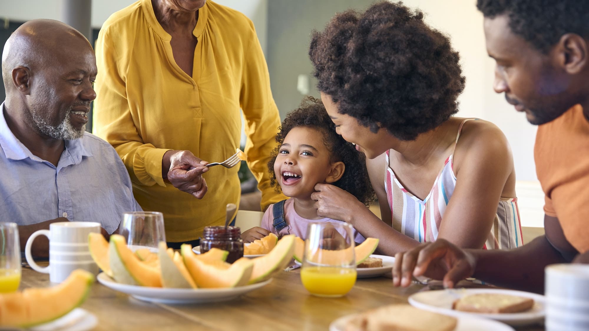 Family Shot With Grandparents Parents And Granddaughter Eating Breakfast Around Table At Home