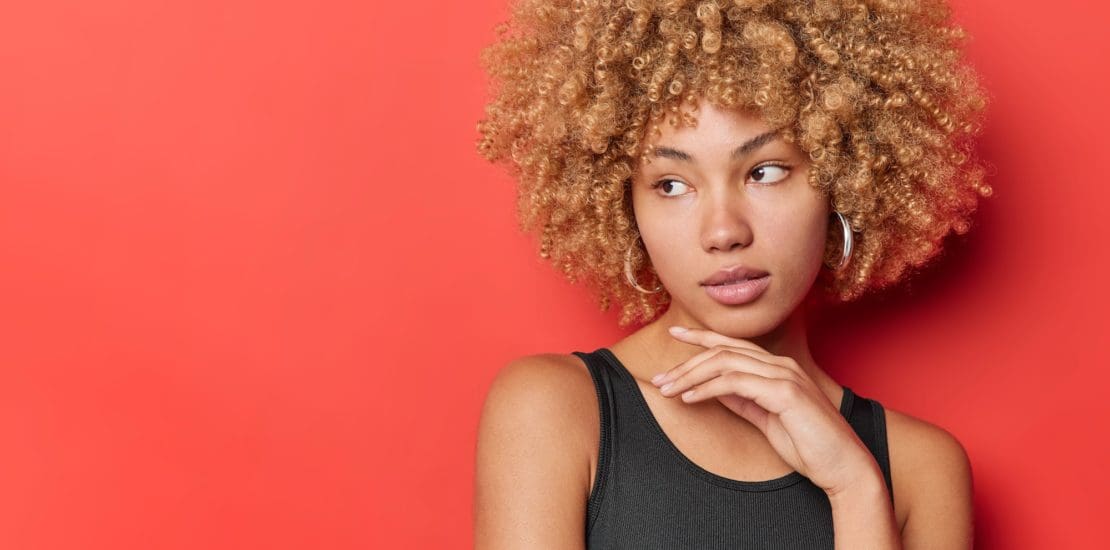 Pensive curly haired woman with natural beauty touches jawline concentrated aside dressed in black t