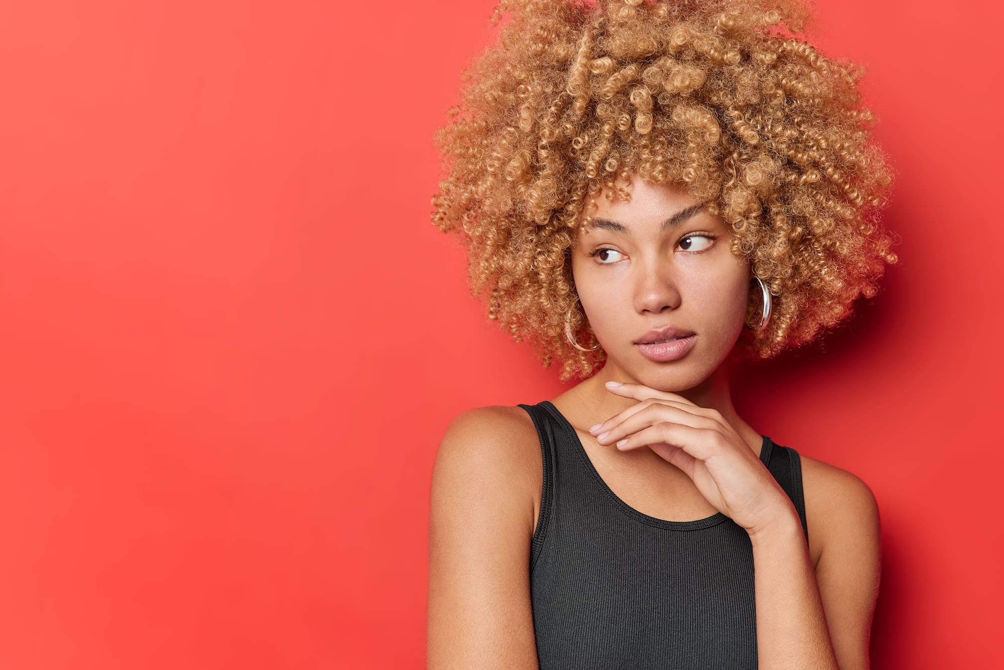 Pensive curly haired woman with natural beauty touches jawline concentrated aside dressed in black t