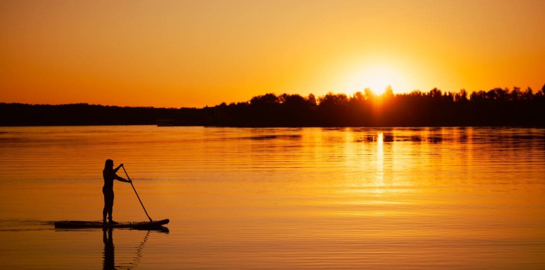 Silhouette of woman paddling oar on sap board at orange sunset on lake in summer, leisure activity