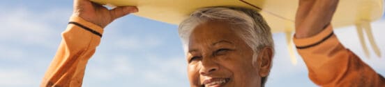 Happy biracial senior woman carrying yellow surfboard over head at beach during sunny day