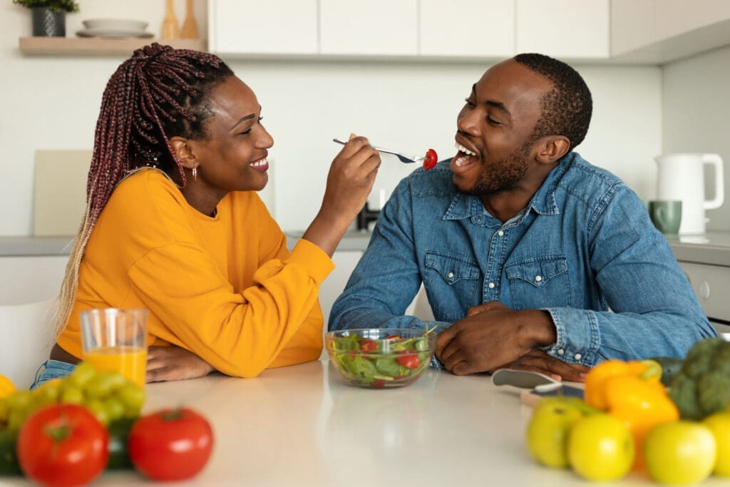 Romantic african american couple eating healthy salad in kitchen, happy wife feeding her husband