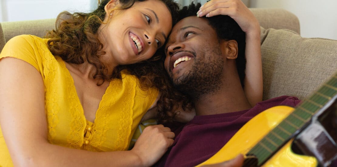 Smiling young multiracial couple romancing while lying with guitar at home