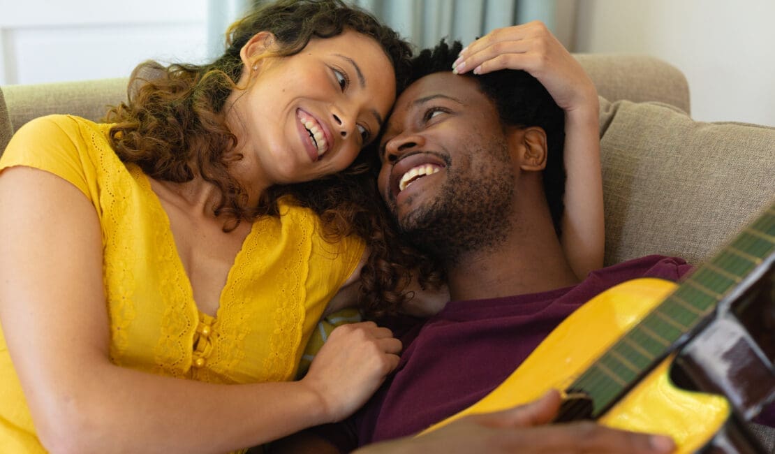 Smiling young multiracial couple romancing while lying with guitar at home