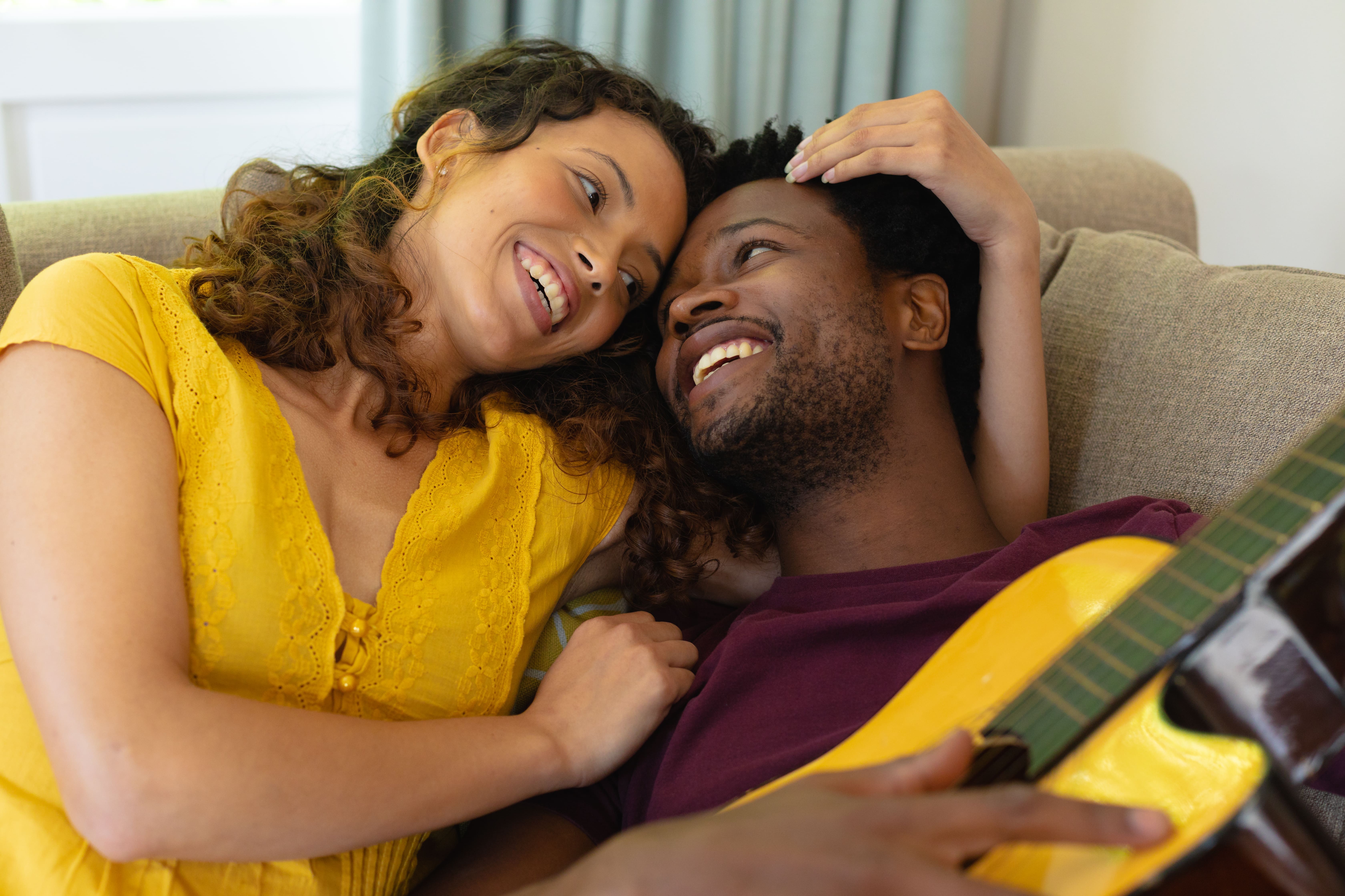 Smiling young multiracial couple romancing while lying with guitar at home