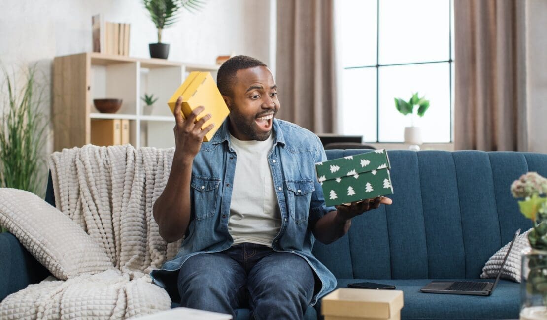 Excited african guy opening gift boxes on couch